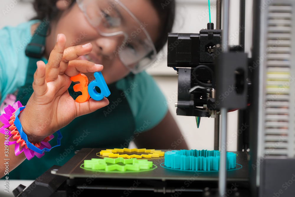 A smart latino student wearing multi-colored 3d printed shapes as jewelry, next to a 3d printer with designs on the heated print bed, and holding 3d letters in her hand.